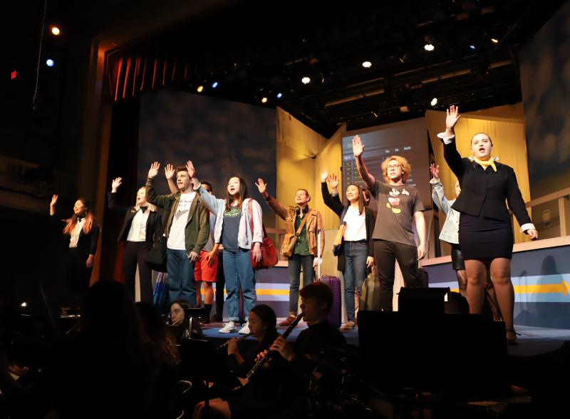 Actors on stage raising their hands singing, with musicians below
