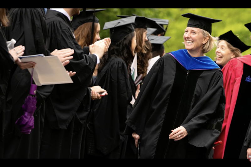 FAS dean Hopi Hoekstra, wearing a graduation cap and gown, smiles as she walks alongside an applauding group of cap- and gown-wearing graduates.