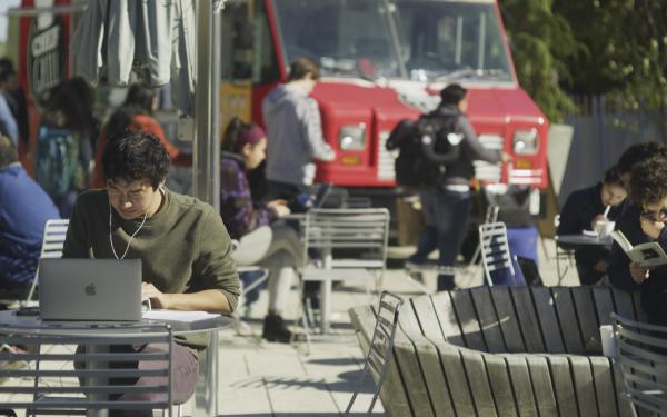 A student in a green sweater sits at a laptop at an outside table. Students in the background are getting lunch from a foodtruck.