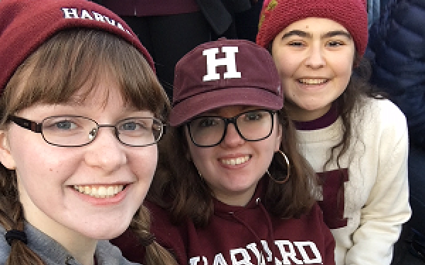 Three girls in Harvard gear take a selfie in the bleachers of a football stadium.