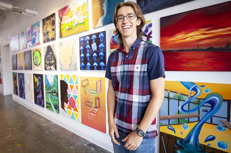 First-year student, Ezra Feder, standing in front of paintings and smiling.