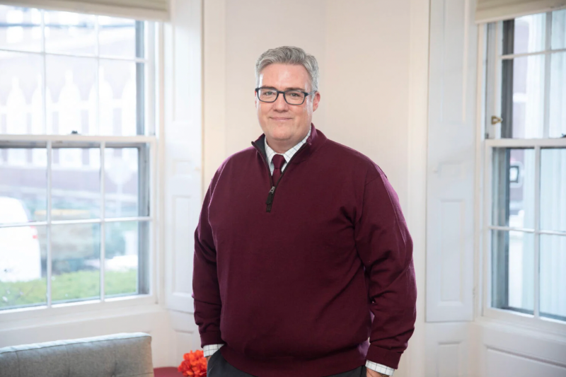 Image of the new Dean of Students, Thomas Dunne, standing in front of windows in the corner of a room. 