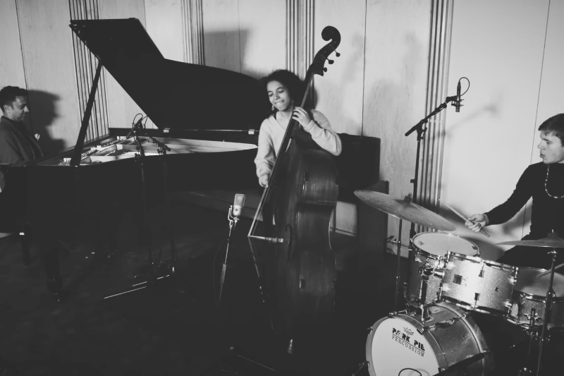 A black and white picture of a pianist playing the piano on the left, a bassist playing the bass in the middle, and a drummer playing the drums on the right.