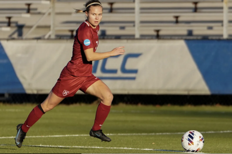 Harvard soccer player Josefine Hasbo, wearing the Harvard team's crimson uniform, dribbles the ball up the field. Some bleachers and a banner with the ACC logo lie in the background.