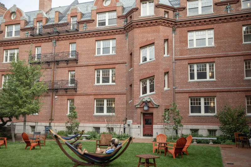 Image of a brick building with windows known as Randolph Hall, with two people sitting in a Hammock outside on a green lawn. 