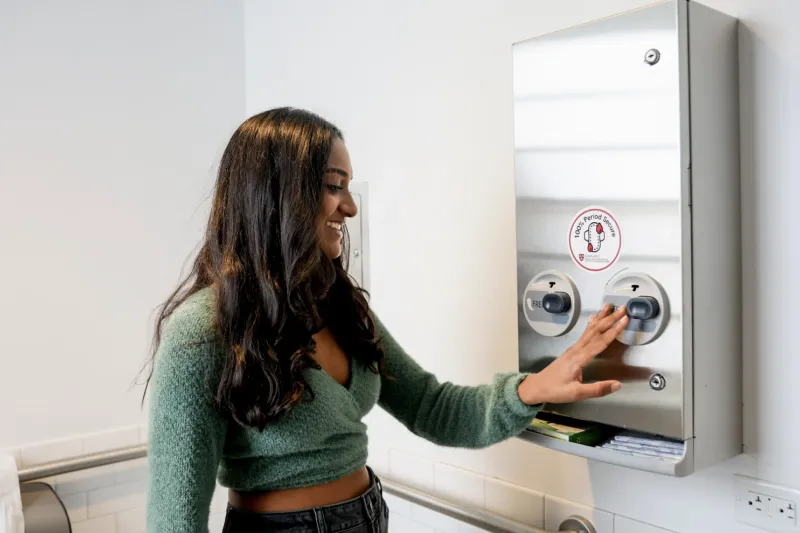 Girl standing in front of a menstrual product dispenser. 