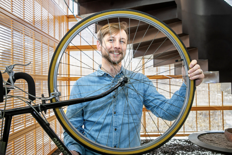 Cory Gills standing and holding up a bike wheel in front of his face. 