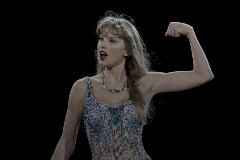 Taylor swift standing in a sparkly body suite with her left arms held up flexing her bicep. 