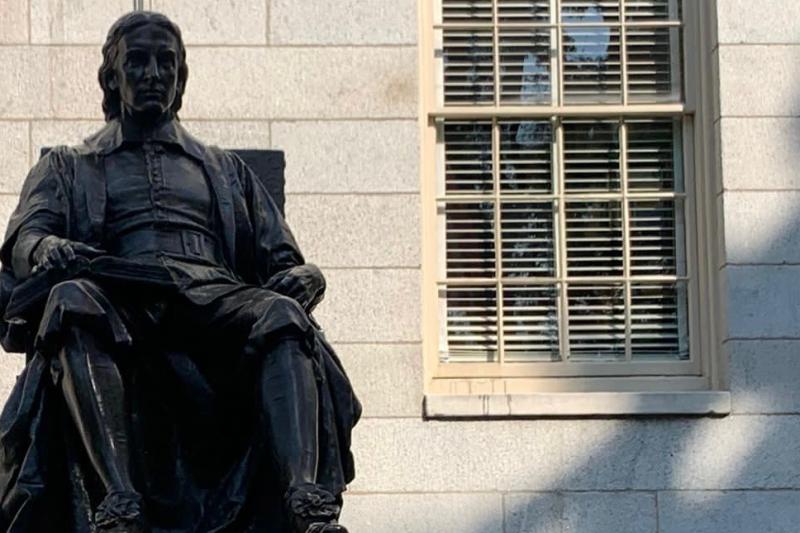 This is a picture of the top of the John Harvard Statue, which shows only the shape of the statue itself, and not the base with it's description. The statue shows a man posing in a sitting, very academic looking, position. 