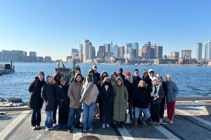 A group of people in winter clothing standing on a waterfront with a city skyline in the background under a clear blue sky.