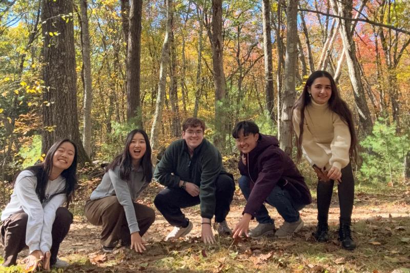 A group of students crouch and smile while touching the ground of Harvard Forest.