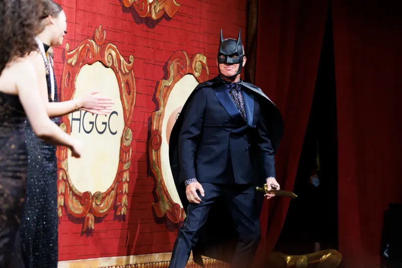 Person in a Batman mask and cape stands on stage in a suit, with a red curtain and another person clapping.
