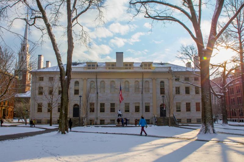 Historic campus building with an American flag and statue in front, surrounded by snow and tall trees with people walking.