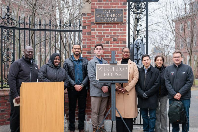 Group of people stand smiling by the Winthrop House gate and sign during an outdoor ceremony.