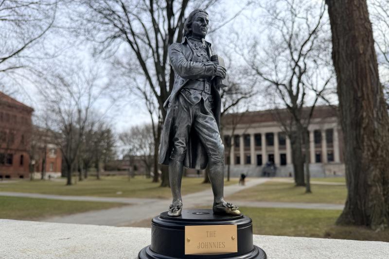 Small bronze trophy of a colonial-era man labeled “The Johnnies” sits on a ledge in a tree-lined college quad with a large library in the background.
