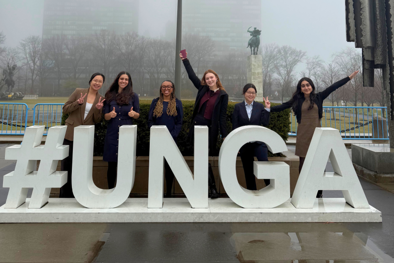 Six people pose smiling behind large “#UNGA” letters outside on a foggy day.