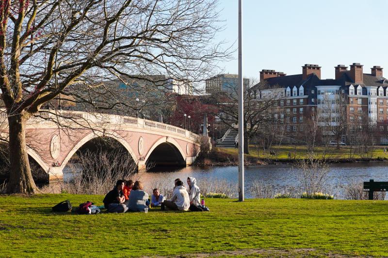 Students relax on a grassy riverbank near a stone bridge, with brick dorm buildings across the water.