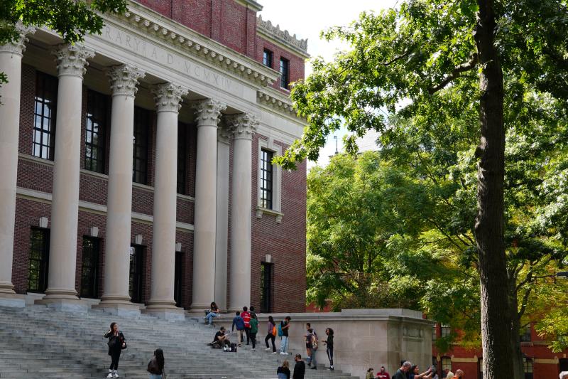 Students and visitors gather on the large front steps of Harvard's brick and columned Widener Library on a sunny day with lush green trees.