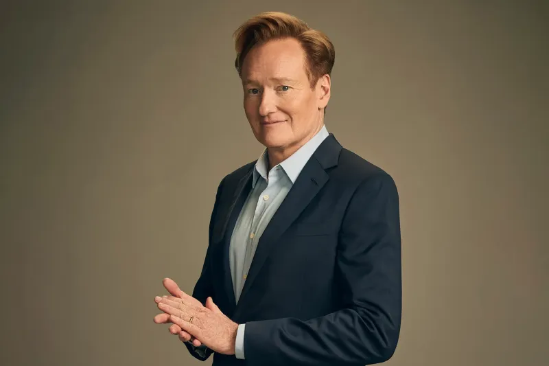 Conan O'Brien, in suit posing behind a concrete colored background.