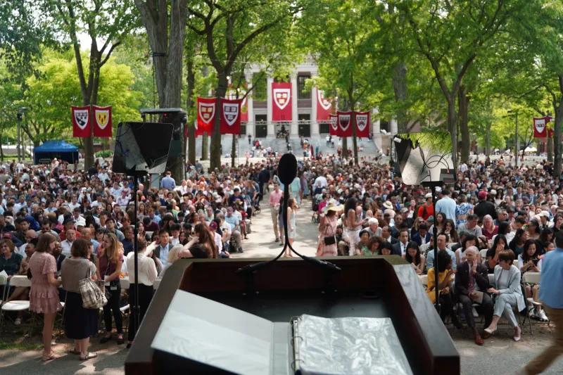 View from the podium toward a large crowd seatd in Harvard Yard, framed by trees and crimson banners.