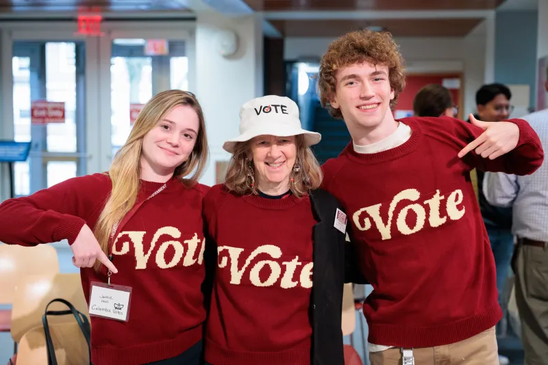 Two students (left and right) and a woman (middle) wearing matching red sweatshirts that say "Vote".