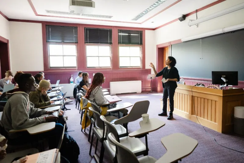 A female professor standing in the front of the classroom teaching to a room of students seated at their desks.
