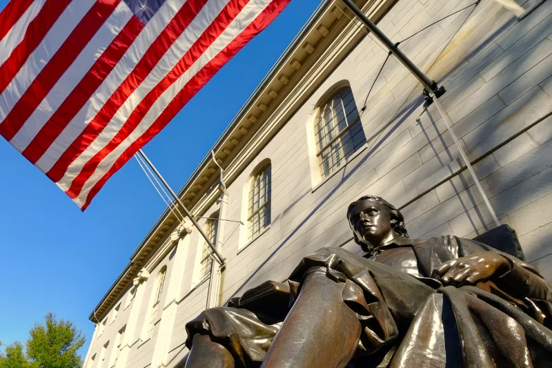 John Harvard statue outside a stone campus building beneath an American flag on a clear day.