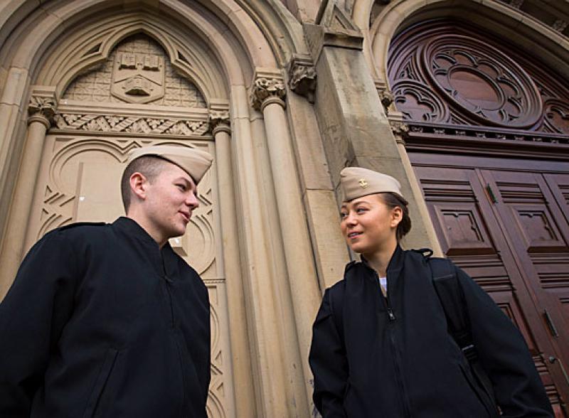 Two students in dark jackets and tan hats stand in front of a Harvard Chapel door