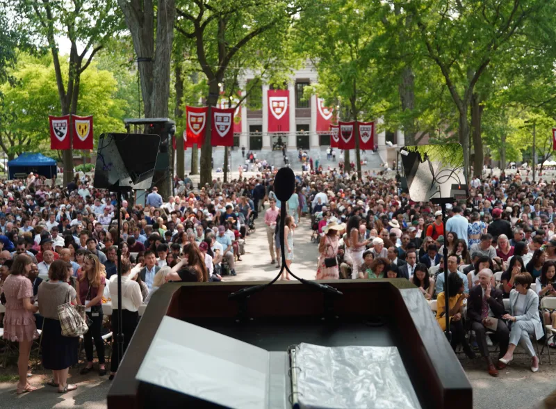 View from the podium toward a large crowd seatd in Harvard Yard, framed by trees and crimson banners.