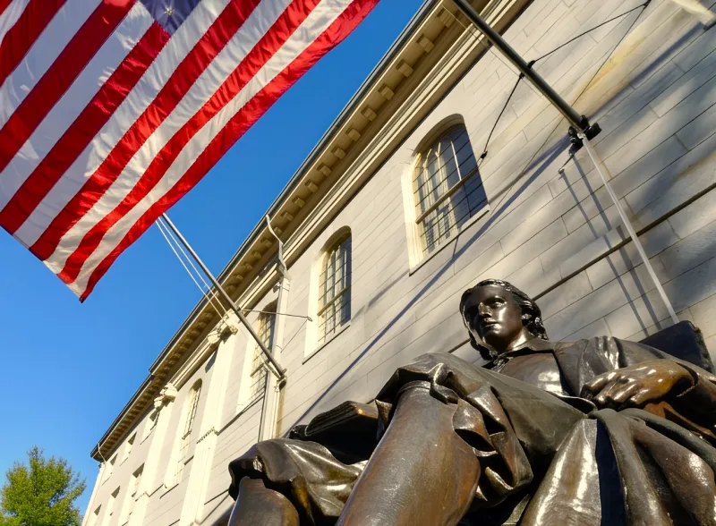 John Harvard statue outside a stone campus building beneath an American flag on a clear day.