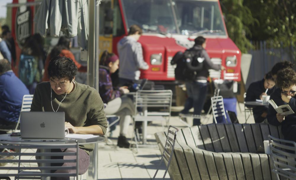 A student in a green sweater sits at a laptop at an outside table. Students in the background are getting lunch from a foodtruck.