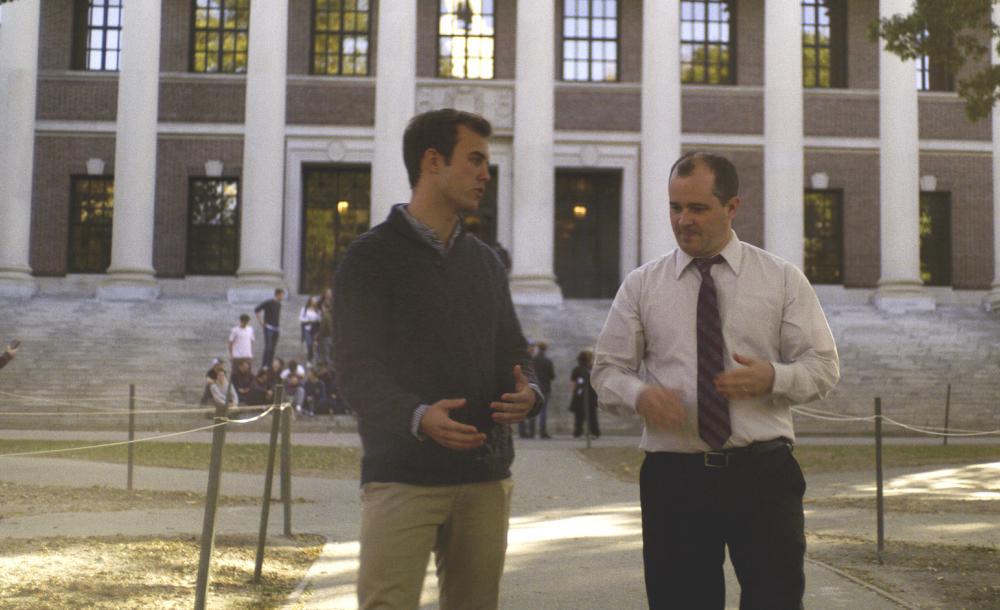 A student and a faculty walk and talk through Harvard Yard.