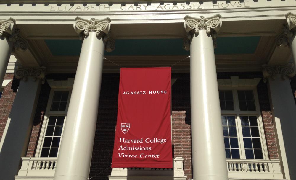 A picture of Agassiz House, taken during my first visit to Harvard. There is a big red banner that hangs between the white columns of the building and reads,"Agassiz House Harvard College Admissions Visitor Center." 