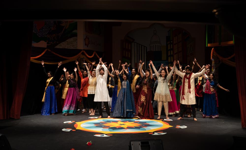 Students wearing South Asian clothing all preparing to bow on stage in the finale of Harvard Ghungroo 2020, with a Rangoli set in front of them.