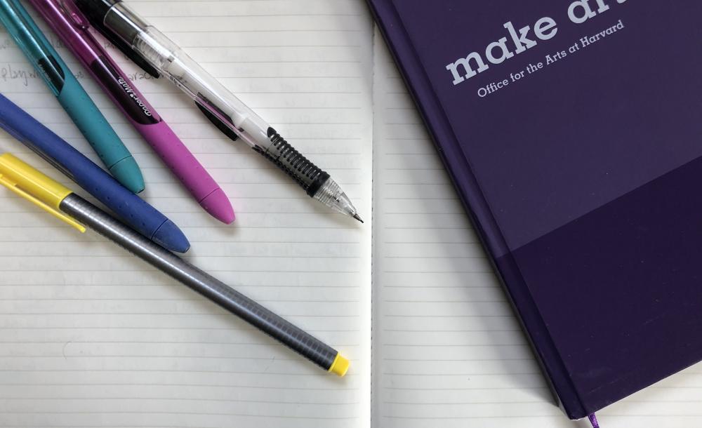 An open notebook. On top there is another purple notebook that says "Make Art" on the cover, along with a variety of pens and pencils.
