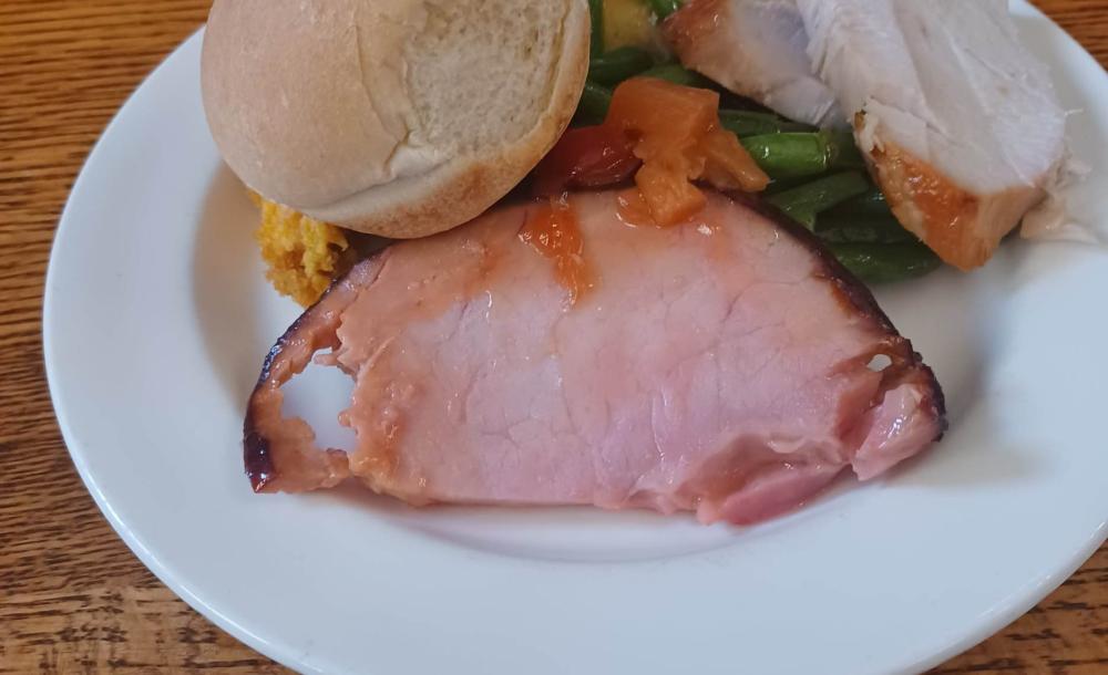 White plate piled with a slice of ham, turkey, a bread roll, green beans, and sweet yams.