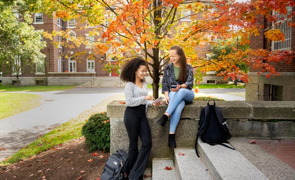 Two students stand on staircase with fall trees behind them