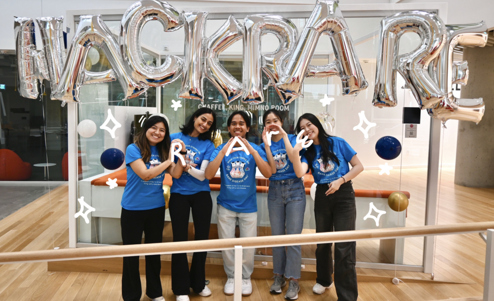 A group of five students wearing blue shirts in front of silver balloons that read &quot;HACK RARE.&quot; They are making symbols with their hands to spell out &quot;RARE.&quot;