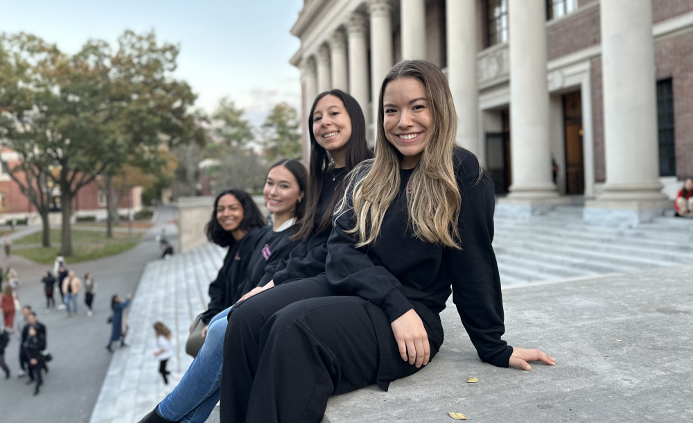 Four people seated on an outdoor marble platform in front of a large building with columns, smiling at the camera. Trees and people walking are visible in the background.