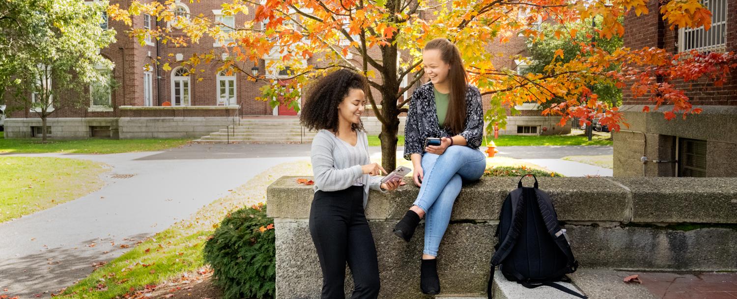 Two students stand on staircase with fall trees behind them