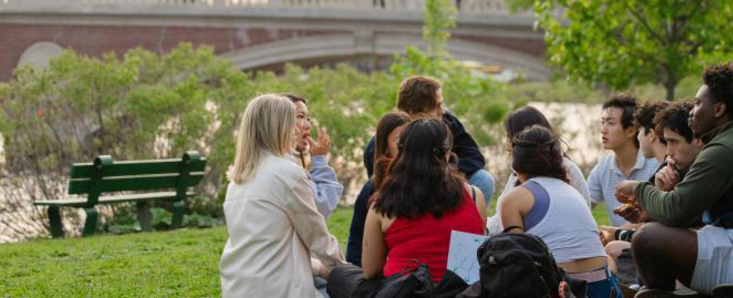 students sitting in a circle on the ground next ro the charles river