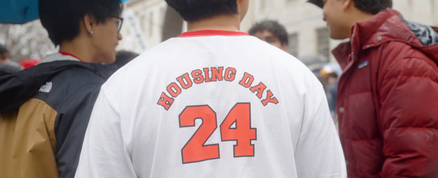 Back of a T-shirt with "Housing Day 24" in red letters, worn in a crowd.