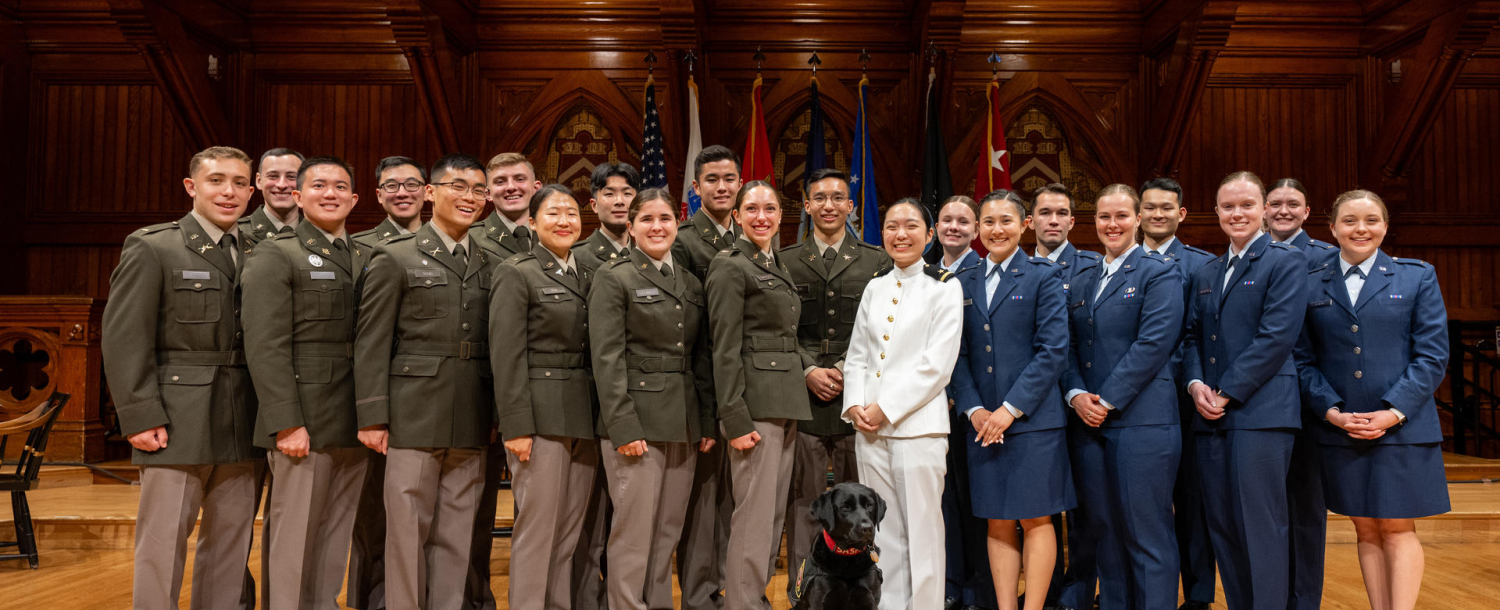 A group of ROTC cadets in uniform pose for a photo indoors with flags in the background.