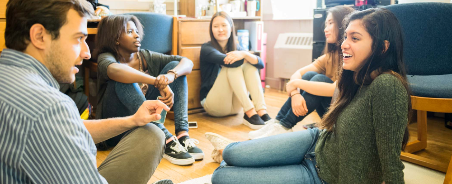 Group of college students sit on a dorm room floor talking together.