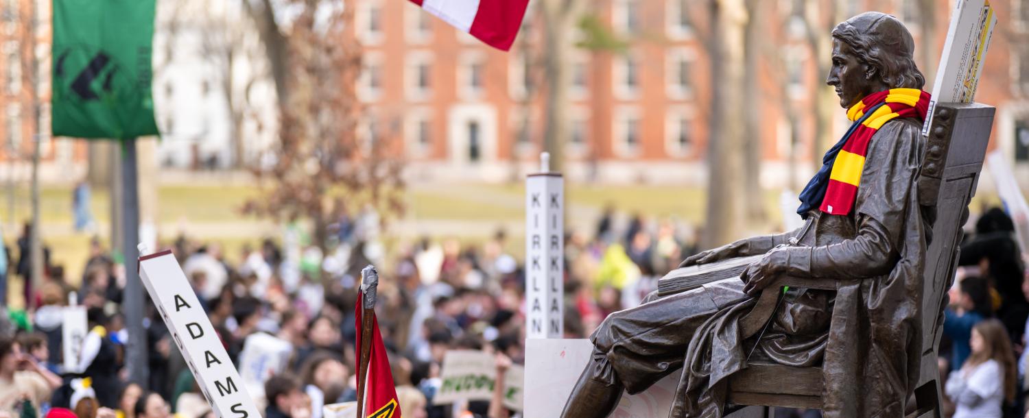 Bronze seated statue wearing a colorful scarf overlooks a large crowd of students holding college house banners and flags.