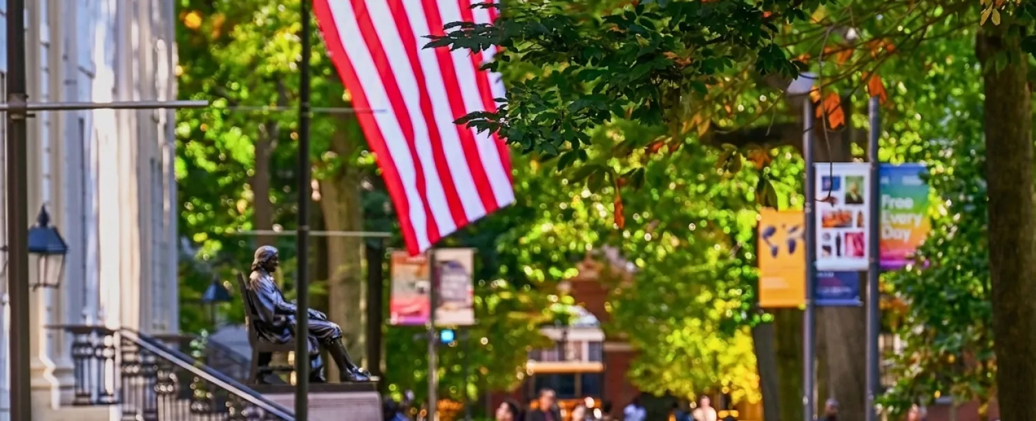 Students walk along a tree-lined campus path beneath an American flag and a seated statue.