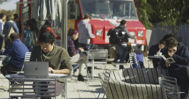 A student in a green sweater sits at a laptop at an outside table. Students in the background are getting lunch from a foodtruck.