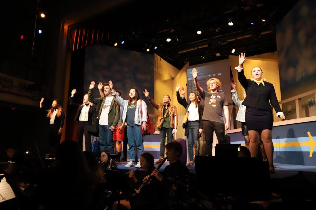 Actors on stage raising their hands singing, with musicians below