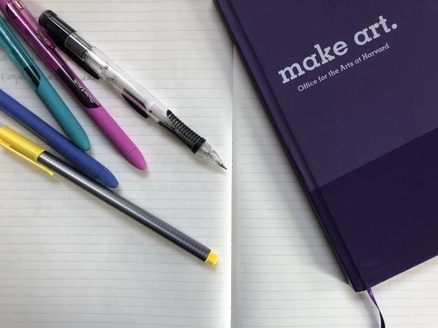 An open notebook. On top there is another purple notebook that says "Make Art" on the cover, along with a variety of pens and pencils.