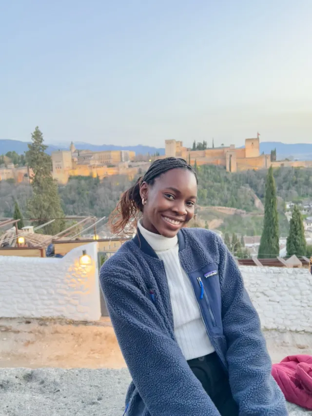 Writer smiling in front of Granada's Alhambra, a castle-like structure on the horizon.